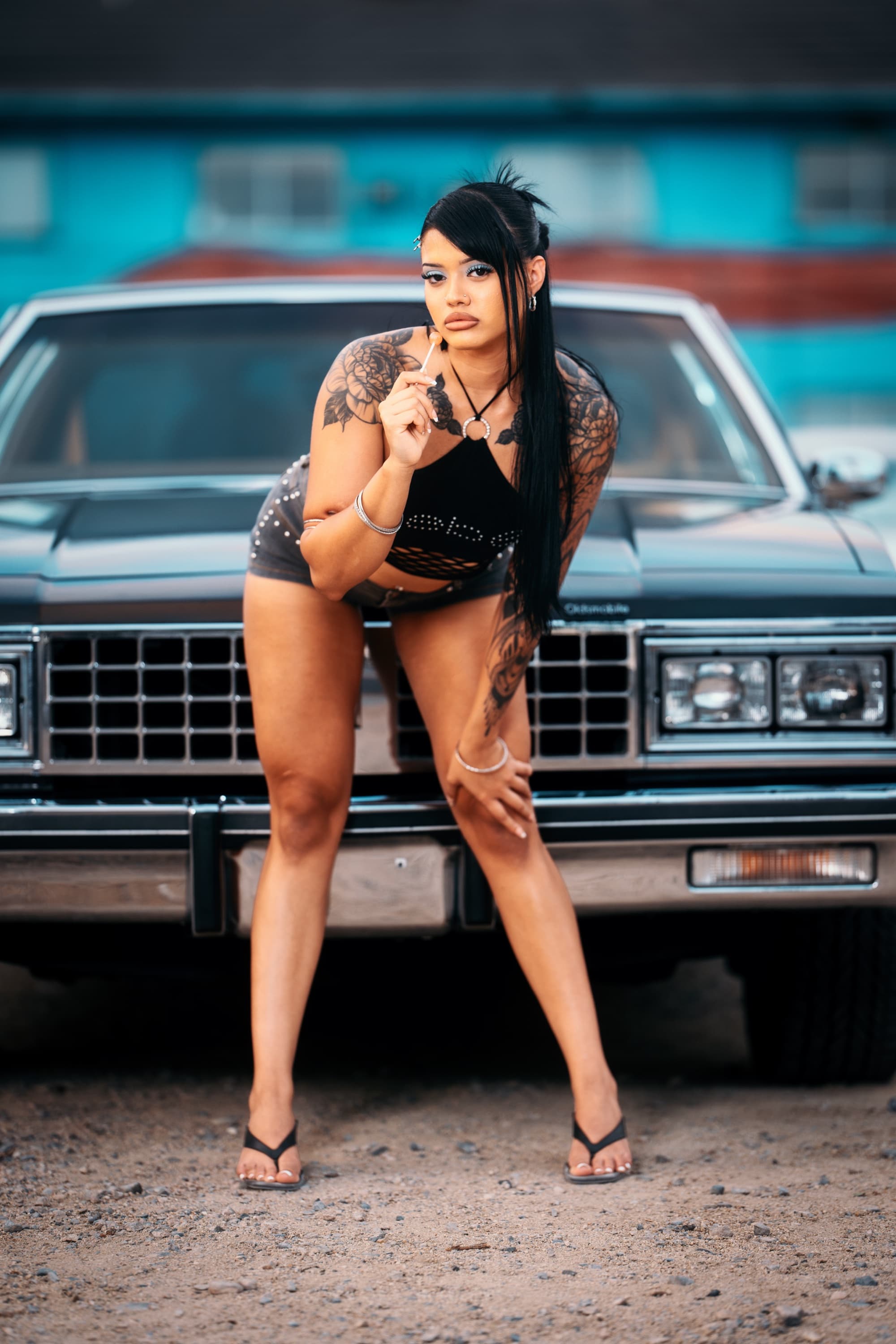 Model leaning on a dark Oldsmobile lowrider in front of a bright teal building