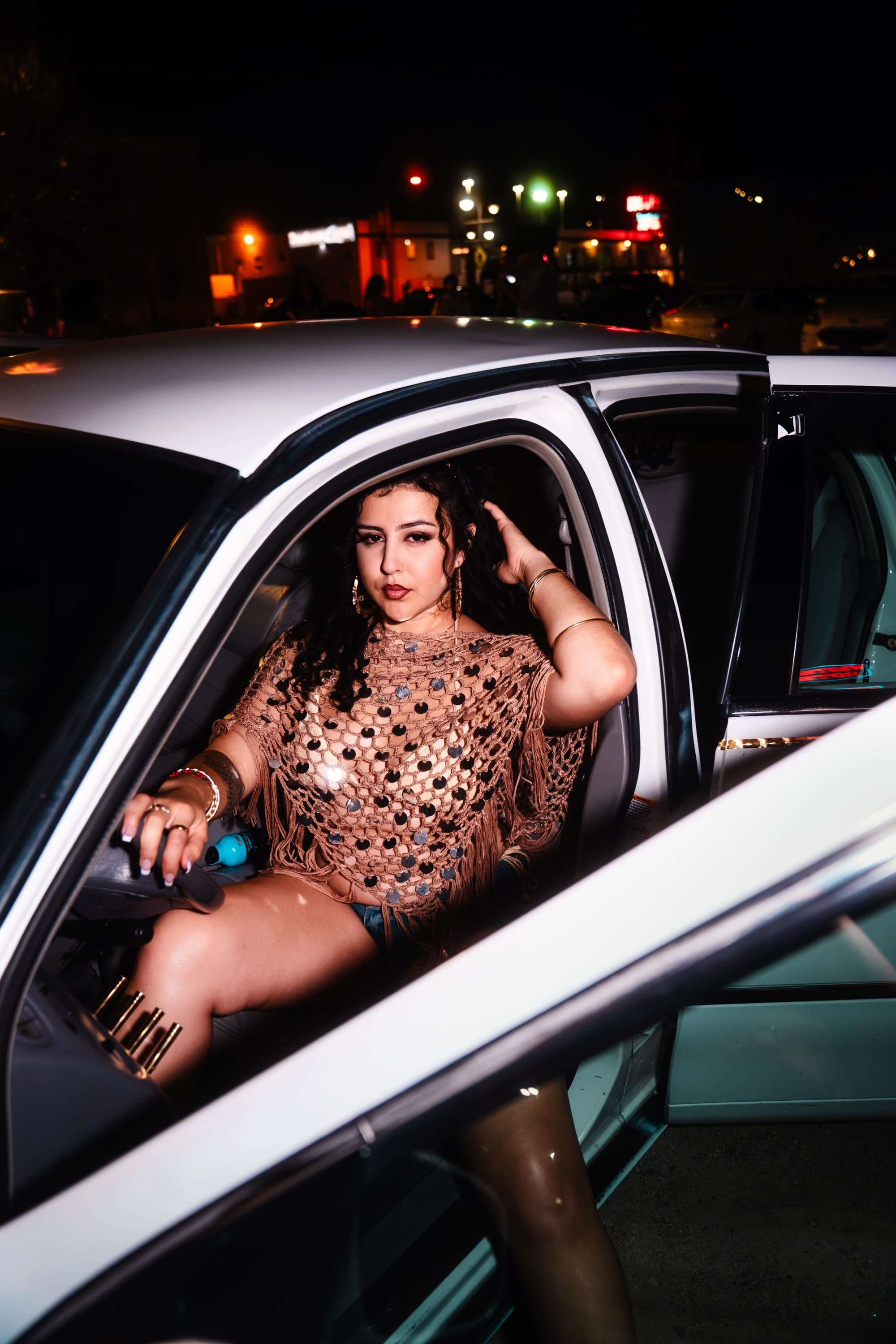 Model in the driver's seat of a white lowrider at night with the door open under city lights