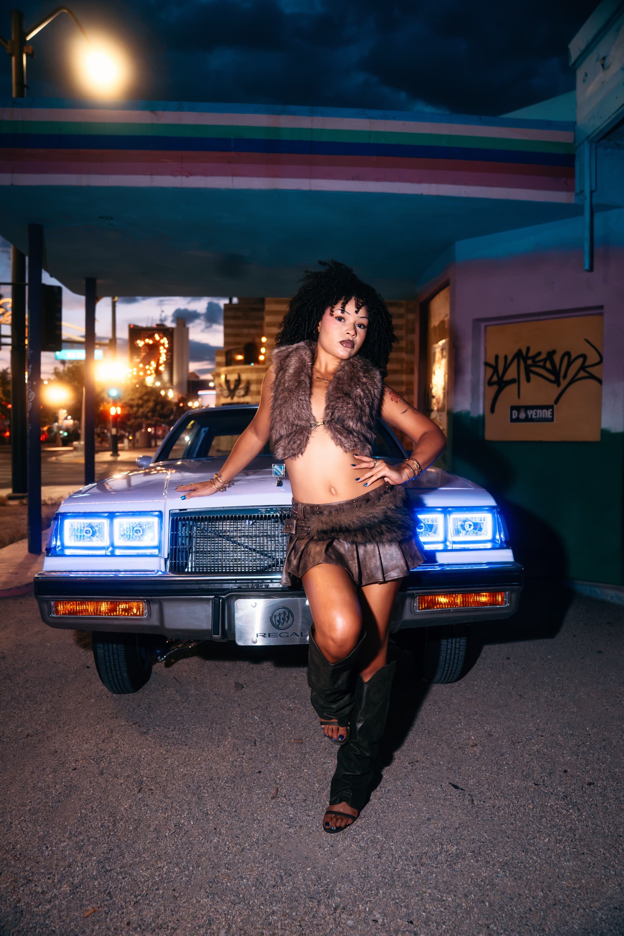 Model in a fur vest in front of a white Buick Regal with glowing blue halo headlights at dusk