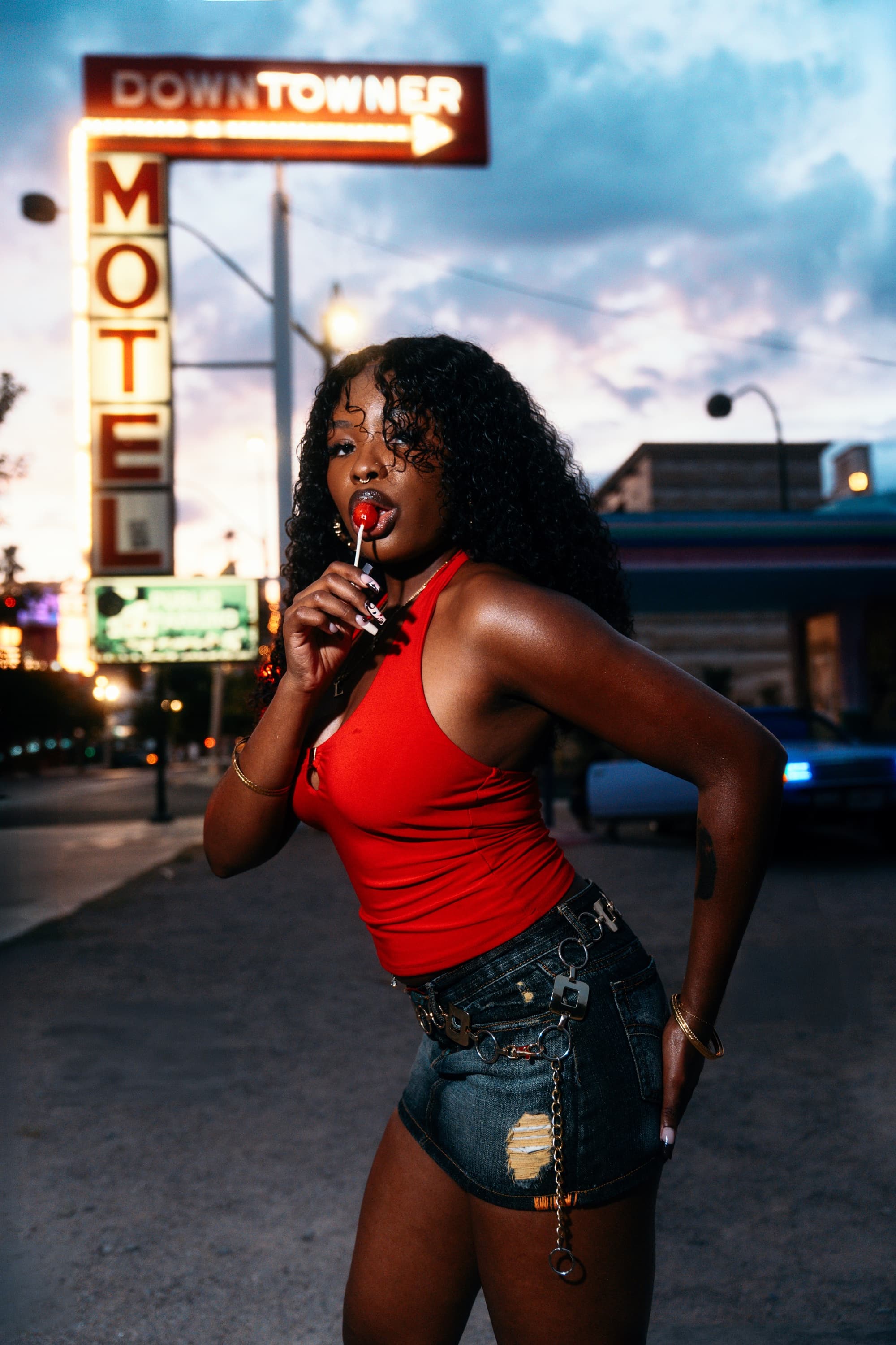 Model in a red top with a cherry lollipop under the glowing Downtowner Motel neon sign at dusk