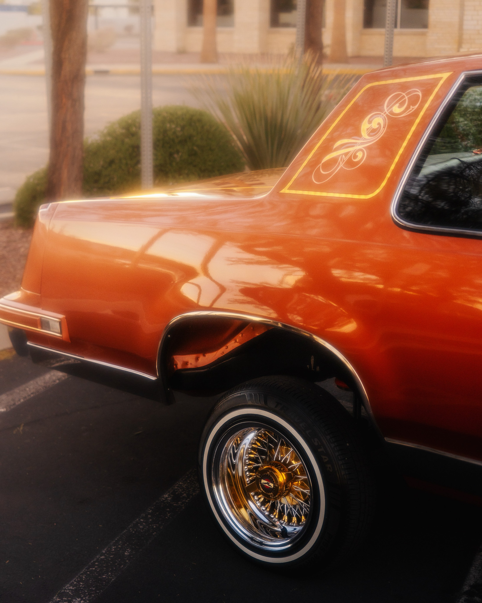 Orange Cutlass lowrider detail shot with gold wire wheels and pinstripe art